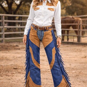 May include: A woman in a white shirt with brown suede details, a straw cowboy hat, and blue and brown fringed chaps. She wears a brown belt and boots. The background shows a rural setting with a fence and trees.