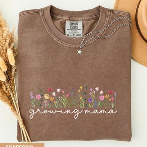 Puede incluir: Una camiseta de cuello redondo marrón con acabado lavado vintage. El frente presenta un gráfico colorido de flores silvestres sobre el texto cursivo blanco "growing mama". Un collar con colgante de estrella plateada descansa en el cuello y la etiqueta Comfort Colors es visible.