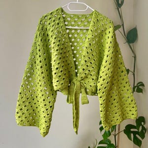 May include: A lime green crochet wrap top featuring an open shell stitch pattern, wide bell sleeves, and a front tie closure. The garment is constructed with a lightweight, airy yarn texture and is displayed on a white plastic clothes hanger.