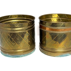 May include: Two cylindrical brass planters featuring a textured diamond pattern etched into the metal surface. Each container has a smooth horizontal band near the top rim and a clear glass base, showcasing a metallic gold finish throughout.