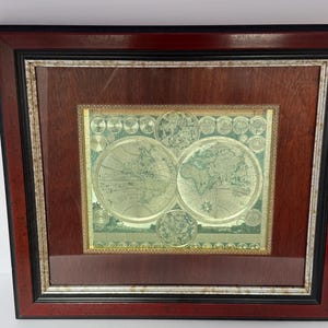 May include: Framed vintage world map print. The map is a detailed illustration with a gold and green color scheme. The frame is a dark wood with a black and gold inner border, adding a classic touch to the artwork.
