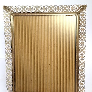 Puede incluir: Un marco de fotos rectangular dorado con un diseño calado ornamentado. El marco presenta un borde decorativo con motivos de corazón y volutas. La parte trasera es de cartón corrugado. El marco está diseñado para contener una fotografía o una obra de arte.