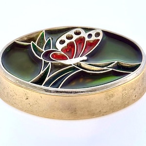 May include: An oval-shaped metal brooch with a gold-toned finish and a raised edge. The center features a decorative inlay with a butterfly motif in red and white, set against a dark green background with stylized leaf accents in silver.