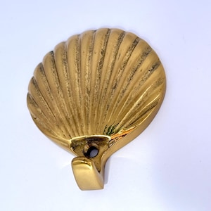 May include: A brass seashell-shaped wall hook. The hook is gold-colored and has a detailed, textured surface. The hook is designed to be mounted on a wall and used for hanging items. The seashell design adds a decorative touch.