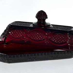 Puede incluir: Una mantequera de vidrio rojo rubí con base rectangular y tapa abovedada. El plato presenta intrincados patrones en relieve y un pomo texturizado en la parte superior. Un diseño clásico de estilo vintage.