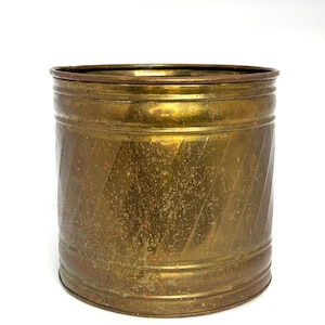 May include: A cylindrical brass container featuring a weathered, tarnished patina and a series of diagonal, embossed stripes wrapping around the exterior. The metal surface displays visible oxidation spots and a warm, aged golden-brown color.