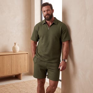 May include: Olive green textured polo shirt with a quarter-zip closure and matching shorts. The outfit has a waffle-like pattern. The model is wearing a watch with a brown leather band.