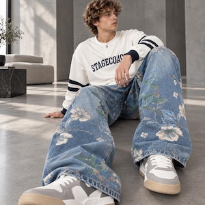 Op de afbeelding: Een wit shirt met lange mouwen, donkerblauwe strepen op de mouwen en de tekst STAGECOACH, gecombineerd met een wijde blauwe spijkerbroek met bloemenprint en wit-grijze sneakers met beige zolen.