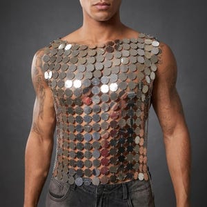 Puede incluir: Un top de cota de malla plateado hecho de discos metálicos circulares. El top sin mangas se sujeta con pequeñas cadenas y cubre el torso. El top se usa sobre la piel desnuda. El fondo es gris liso.