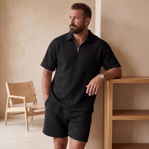 May include: Black textured polo shirt with a silver zipper and matching shorts. The outfit is made of a waffle-like fabric. The model is wearing a silver watch. The background is a neutral beige.