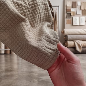 May include: Close-up of a beige quilted fabric with a diamond pattern. The fabric is held up, showing the texture and stitching. The background includes rolls of fabric and a display of fabric swatches.