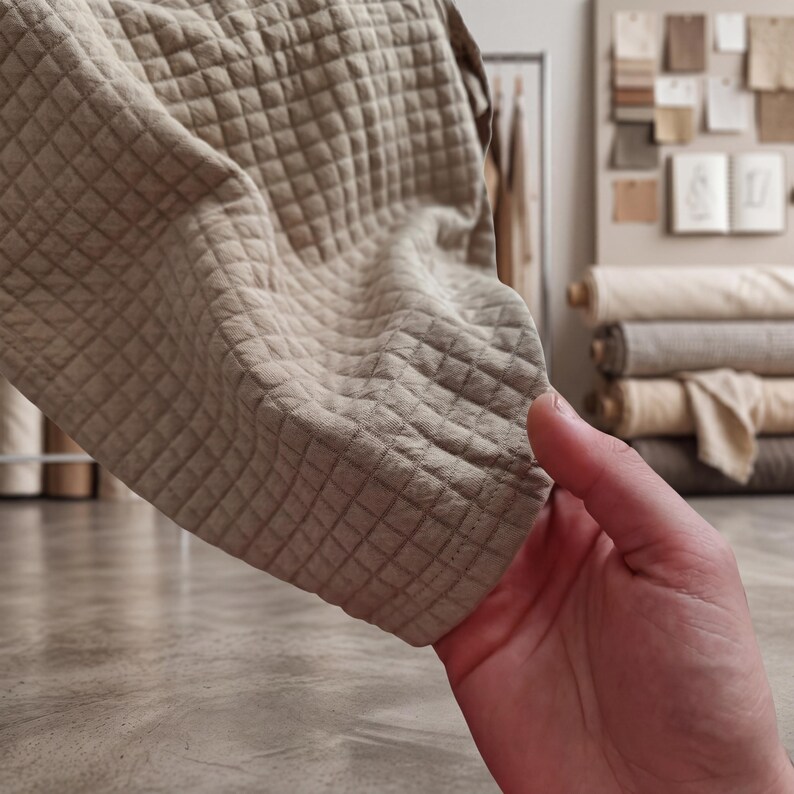 May include: Close-up of a beige quilted fabric with a diamond pattern. The fabric is held up, showing the texture and stitching. The background includes rolls of fabric and a display of fabric swatches.