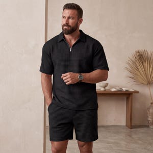 May include: Black textured polo shirt with a silver zipper and matching shorts. The shirt has short sleeves and a collared neckline. The shorts have pockets. The outfit is made of a waffle-like fabric.