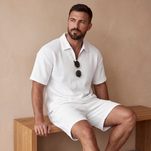 May include: White textured polo shirt with a zipper and matching shorts. The outfit includes sunglasses hanging from the shirt. The clothing has a waffle-like texture. The person is sitting on a wooden bench.