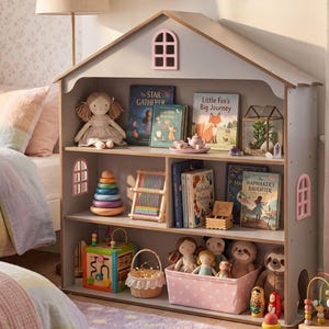 Könnte beinhalten: Ein graues Bücherregal aus Holz in Form eines Hauses, gefüllt mit Kinderbüchern, Spielzeug und Puppen. Die Regale zeigen eine Vielzahl von Gegenständen, darunter ein Holz-Stapelspielzeug, ein Webstuhl und ein rosa gepunkteter Korb. Das Bücherregal hat rosa Fenster- und Türausschnitte.