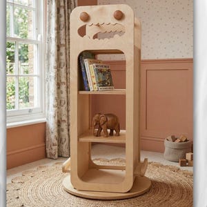 May include: A light-colored wooden bookshelf with a unique design, featuring rounded edges and a playful cutout at the top. The bookshelf has multiple shelves holding books and a wooden elephant figurine. The bookshelf is on a round, woven rug.