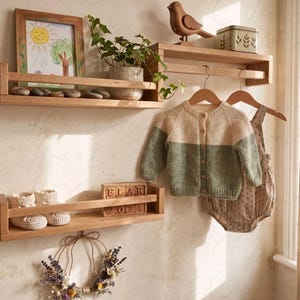 Könnte beinhalten: Zwei Wandregale aus hellem Holz mit vorderen Leisten. Das obere Regal enthält ein gerahmtes Sonnenbild, eine Topfpflanze, einen Holzvogel und eine Dose. Das untere Regal enthält zwei weiße Strickschühchen und Holzklötze mit der Aufschrift ELAR 2024.