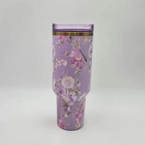 May include: A tall, cylindrical stainless steel tumbler in a light lavender color featuring a repeating floral pattern of pink, white, and purple roses. The drinkware includes a metallic gold rim and a translucent, light purple plastic lid.
