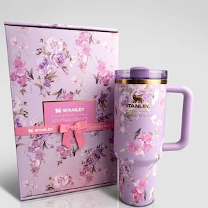 May include: A lavender Stanley tumbler with a floral pattern and a matching gift box. The tumbler features a gold Stanley logo, a purple handle, and a clear lid. The box is decorated with the same floral print, a pink ribbon, and a Stanley LoveShackFancy label.