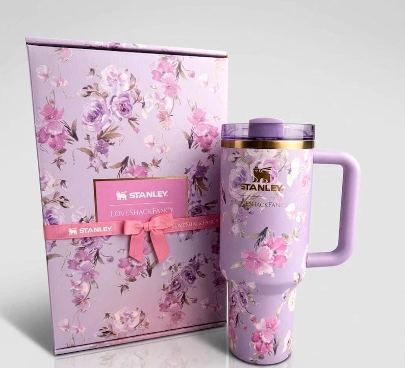 May include: A lavender Stanley tumbler with a floral pattern and a matching gift box. The tumbler features a gold Stanley logo, a purple handle, and a clear lid. The box is decorated with the same floral print, a pink ribbon, and a Stanley LoveShackFancy label.