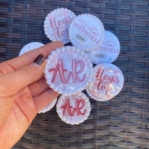 May include: A collection of round, decorative pins with white pearl-like borders. Some pins feature the text "Hogs to" and "AR" in pink script. Other pins have the text "With love from Fayetteville" and a pig illustration.