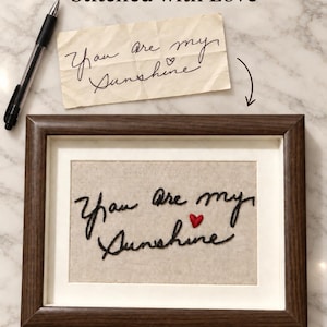 Pode incluir: Uma moldura de madeira escura contendo um painel de tecido bege com o texto bordado em preto "You are my Sunshine" e um pequeno coração vermelho. Acima da moldura, um pedaço de papel exibe o mesmo texto manuscrito, acompanhado por uma caneta preta.