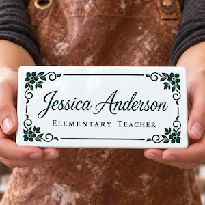May include: A rectangular white ceramic sign featuring dark green floral corner accents and a dark green border. The text reads Jessica Anderson in a script font, with the words ELEMENTARY TEACHER printed in a clean, sans-serif font below.