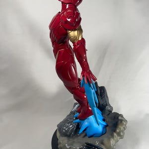 May include: A red and gold Iron Man statue. The figure is in a dynamic pose, with blue energy effects emanating from the feet. The statue is on a rocky base with a black circular stand.