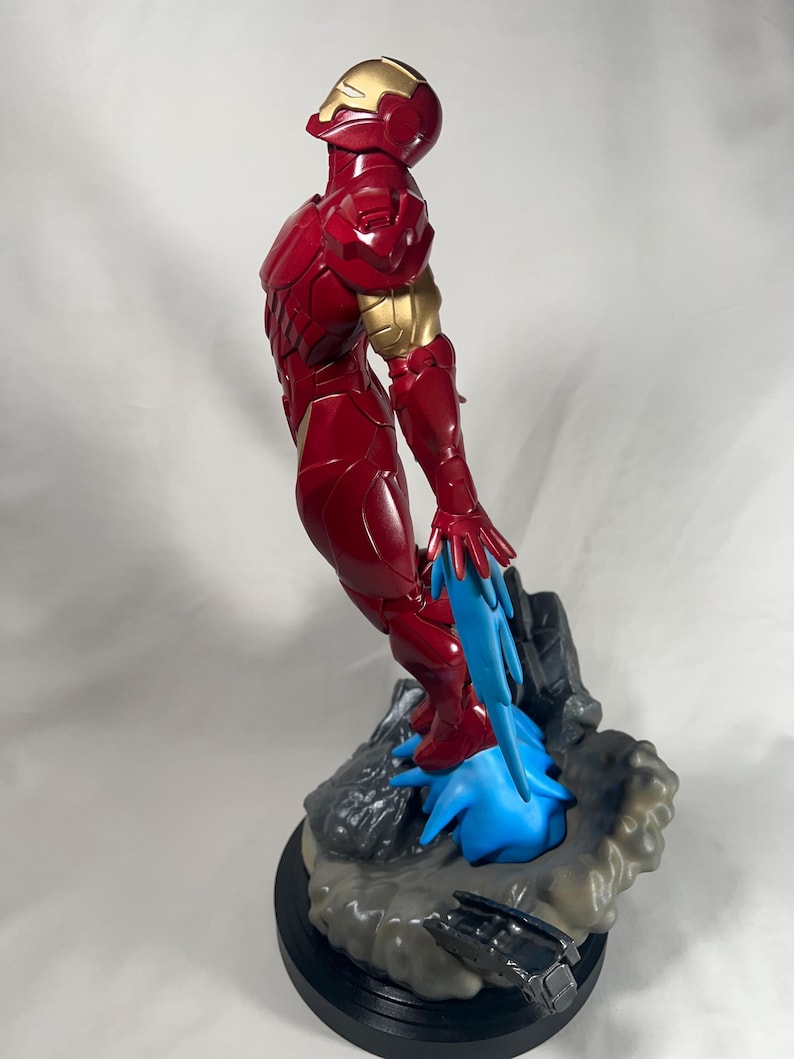 May include: A red and gold Iron Man statue. The figure is in a dynamic pose, with blue energy effects emanating from the feet. The statue is on a rocky base with a black circular stand.