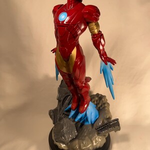 May include: A red and gold Iron Man figurine in a dynamic pose, with blue energy effects emanating from the feet and hands. The figure is on a detailed base with a black circular platform. The helmet and chest feature a glowing blue light.