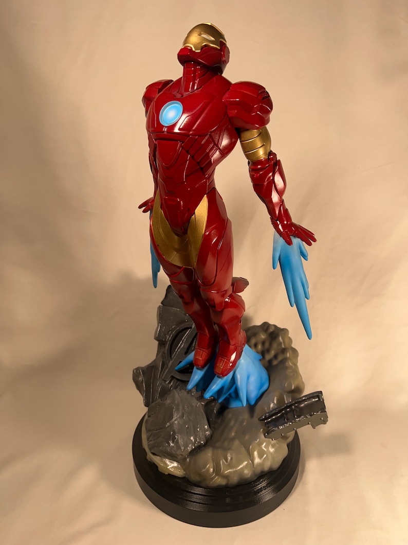 May include: A red and gold Iron Man figurine in a dynamic pose, with blue energy effects emanating from the feet and hands. The figure is on a detailed base with a black circular platform. The helmet and chest feature a glowing blue light.
