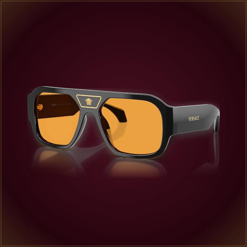 May include: Black sunglasses featuring a thick frame and vibrant orange lenses. A gold-toned metal accent sits on the bridge, displaying the Medusa head logo. The word Versace is printed in gold lettering on the side of the temple arm.