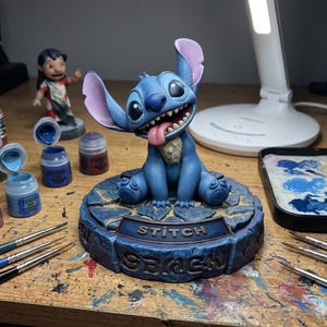 Könnte beinhalten: Blaue Stitch-Figur mit großen Ohren und einem lächelnden Gesichtsausdruck, Zunge heraus. Die Figur sitzt auf einem Sockel mit der Aufschrift "Stitch" und "Beach". Farbzubehör und eine Lilo-Figur befinden sich im Hintergrund.
