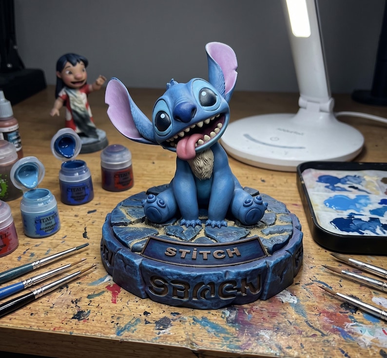 May include: A blue Stitch figurine with large ears and a smiling expression, tongue out. The figurine sits on a base that reads "Stitch" and "Beach". Paint supplies and a Lilo figurine are in the background.