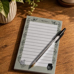 May include: A lined notepad with a pale green border, decorated with floral and butterfly illustrations. The word "NOTES" is at the top. A black pen lies diagonally across the notepad. It rests on a wooden surface.