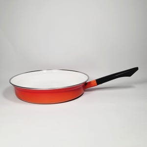 Peut inclure: Poêle à frire en émail orange avec une poignée noire. La poêle a un intérieur blanc.