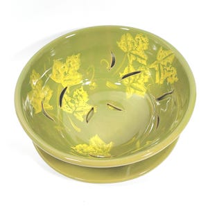 May include: A green ceramic bowl with a matching base. The bowl has yellow leaf designs and cut-out accents. The round bowl has a glossy finish, suitable for serving or display.