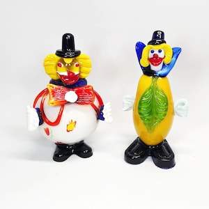 Peut inclure: Deux figurines de clowns en verre. Le clown de gauche a un corps rond blanc avec des accents rouges, des cheveux jaunes, un chapeau noir et un nœud papillon rouge. Le clown de droite a un corps jaune allongé avec une feuille verte, des cheveux jaunes et un chapeau noir.