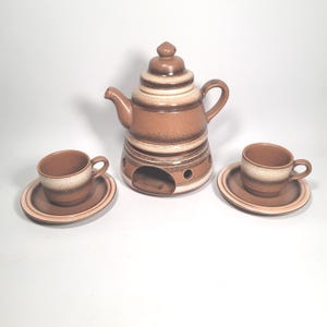 Peut inclure: Un service à thé vintage en céramique dans les tons de marron et de crème. L'ensemble comprend une théière avec un couvercle, deux tasses à thé et deux soucoupes. La théière a un chauffe-plat intégré. Les tasses et les soucoupes ont un design bicolore.