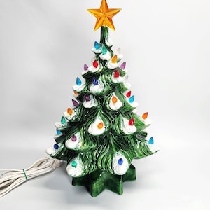 May include: A ceramic Christmas tree with a glossy green finish and white painted snow on the branches. The tree is decorated with small, multi-coloured translucent plastic bulbs and topped with a large, bright orange star-shaped light fixture.