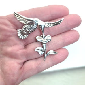 May include: A silver hummingbird brooch, depicted with wings spread, feeding from a flower. The brooch features detailed textures, highlighting the bird's feathers and the flower's petals. The design is intricate and elegant, a piece of jewellery.