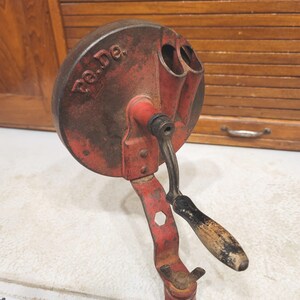 May include: Vintage red and black metal hand-cranked tool with a wooden handle. The tool has a circular top with two openings and the text "Po.De." The handle is black and the metal shows signs of age and wear.