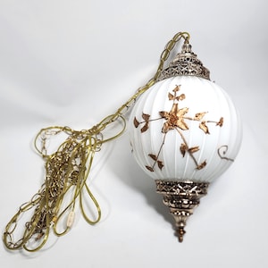 May include: A white, ribbed, spherical glass pendant lamp featuring gold-toned floral and vine motifs. The fixture includes ornate, metallic gold-coloured end caps at the top and bottom, and is suspended by a matching gold-toned metal chain.