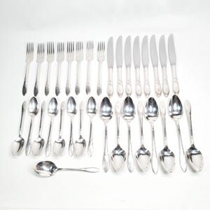 May include: An assortment of silver-coloured cutlery, comprising forks, knives, and spoons, displayed on a white surface. The utensils vary in size and have a reflective finish. The arrangement suggests a formal dining setting.