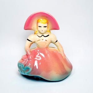 May include: A ceramic figurine of a person wearing a wide-brimmed pink hat and a matching pink dress with a blue accent at the hem. The figure has blonde hair, painted facial features, and a smooth, glossy finish with a gradient colour effect.