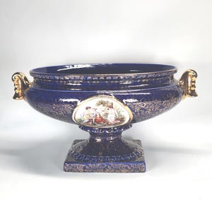May include: A deep blue ceramic bowl with a pedestal base and two gold-toned handles. The exterior features intricate gold floral patterns and a central oval medallion depicting a colourful scene of figures in a landscape setting.