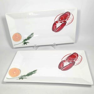 May include: Two rectangular white serving platters. Each platter features a red lobster design with a red border and text. One platter has a slice of orange and a sprig of green herbs. The other platter has the same design.