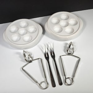May include: Two white ceramic dishes with six indentations each, designed for serving escargots.  Two silver escargot tongs and two silver forks are included.