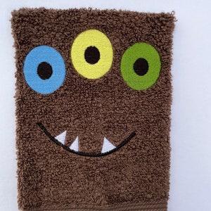 May include: Brown terrycloth hand towel with an embroidered monster face. The monster has three eyes, one blue, one yellow, and one green, and a white toothy grin.