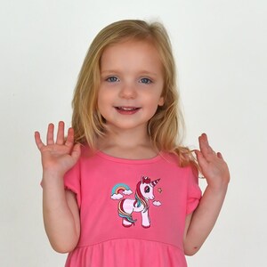 May include: A pink dress featuring a unicorn design with a rainbow and stars. The dress has short sleeves and a round neckline. The unicorn has a pink body, a purple mane, and a golden horn. The girl is smiling and waving.